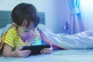 Cute little child watch movie on smartphone at bed. Dangers of blue light can damage eyes. Handsome little boy can be age related macular degeneration from blue light, wear eyeglasses since childhood