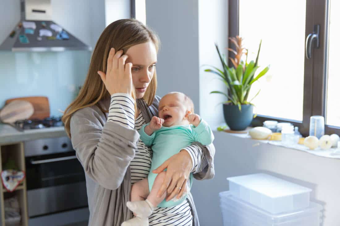 Tired concerned new mother rocking crying baby in kitchen. Portrait of young woman and cute little child in home interior. Motherhood concept
