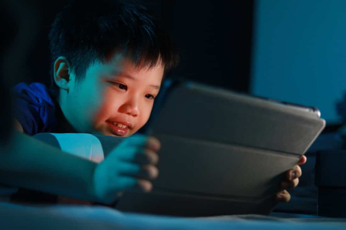 Little Asian boy lying on the bed at night and playing digital tablet. Soft focus image.