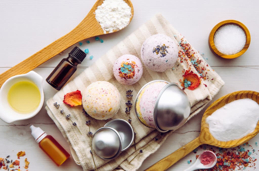 Making bath fizz bombs at home concept. All the ingredients on table on wood spoons: cornstarch, essential oil, dye, citric acid, baking soda, dry herbs, round metal pressing molds.