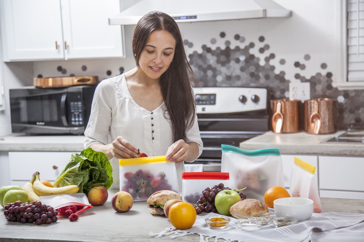 A young mother puts strawberries into a clear food storage bag. She is in the kitchen with a concrete counter top surrounded by other fruits and vegetables that she is preparing and storing.