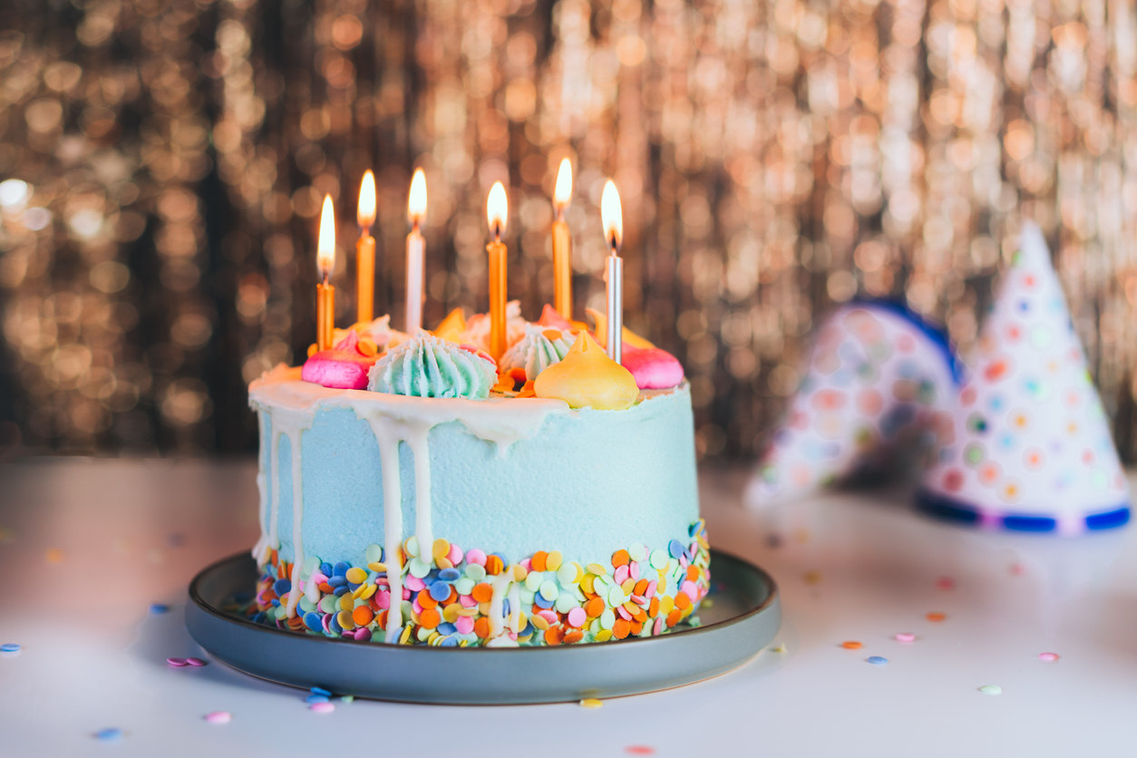 Colorful birthday cake with sprinkles and burning candles and festive caps on the sparkling gold tinsel background. Festive birthday celebration, party. Selective focus, copy space