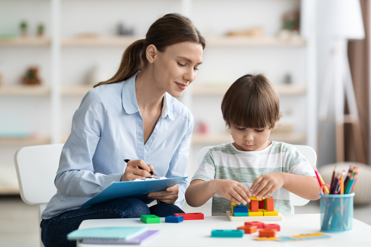 Assessment of kids mental development. Professional woman psychologist watching little boy playing with logical game, evaluating his readiness for preschool, free space