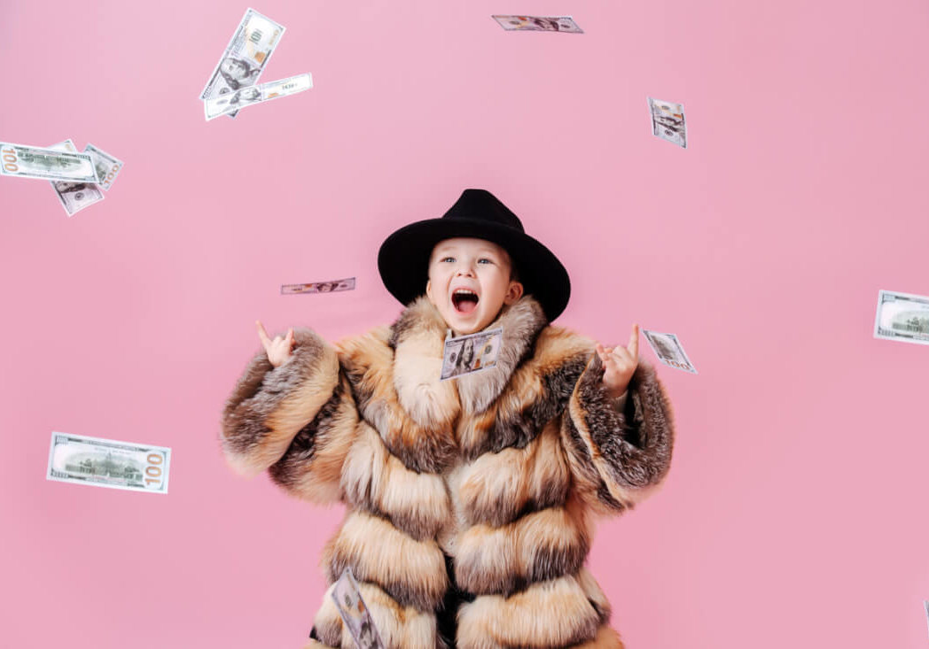Playful little boy in oversized luxurious fur coat and classical hat making rock'n roll gesture under money shower. Over dark pink background.