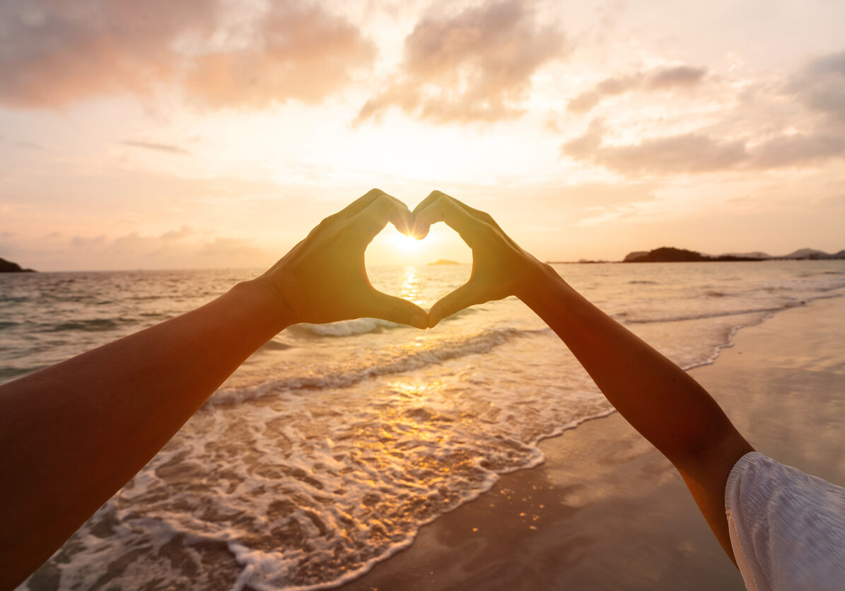 Young couple traveler making heart shape with hands on the beach at sunset, Lovers on honeymoon and Valentine's day concept