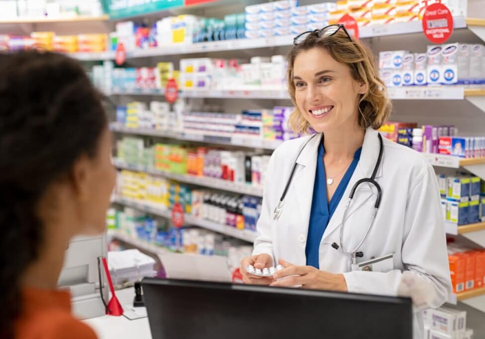 Pharmacist giving medicine to customer in modern pharmacy. Rear view of young woman buying analgesic at pharmacy. Happy doctor giving strip of tablet to customer.