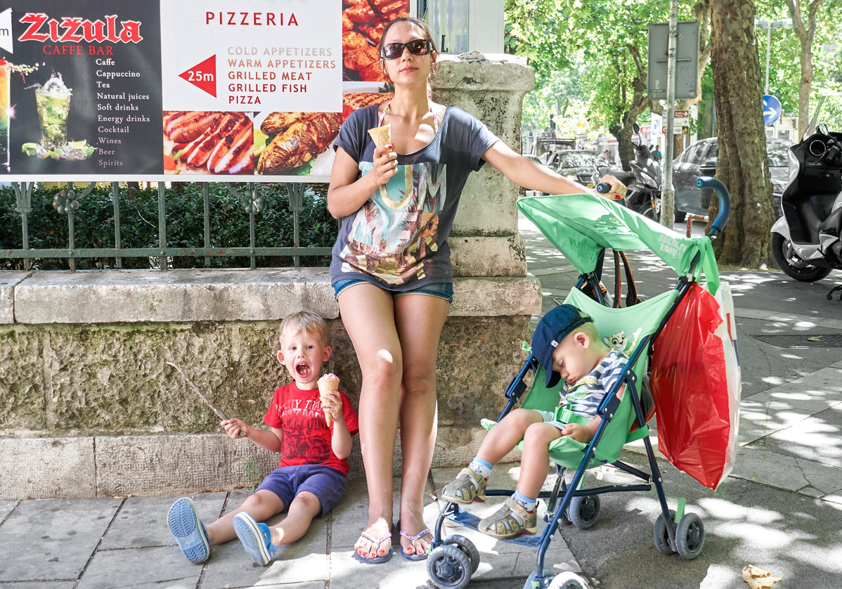 Omis, Croatia - August 13 2013: Mother with stroller and toddler sons eat ice cream standing on street of Omis. Woman in sunglasses and boys enjoy summer vacation in Croatia