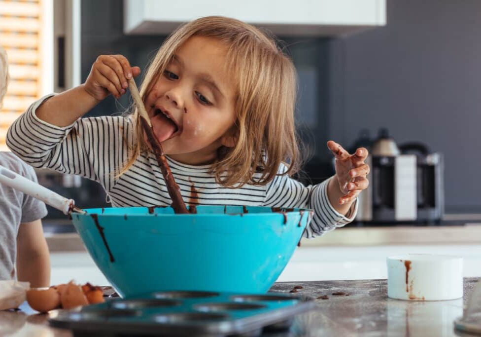 Little girl licking spoon while mixing batter for baking in kitchen  and her brother standing by. Cute little children making batter for baking.